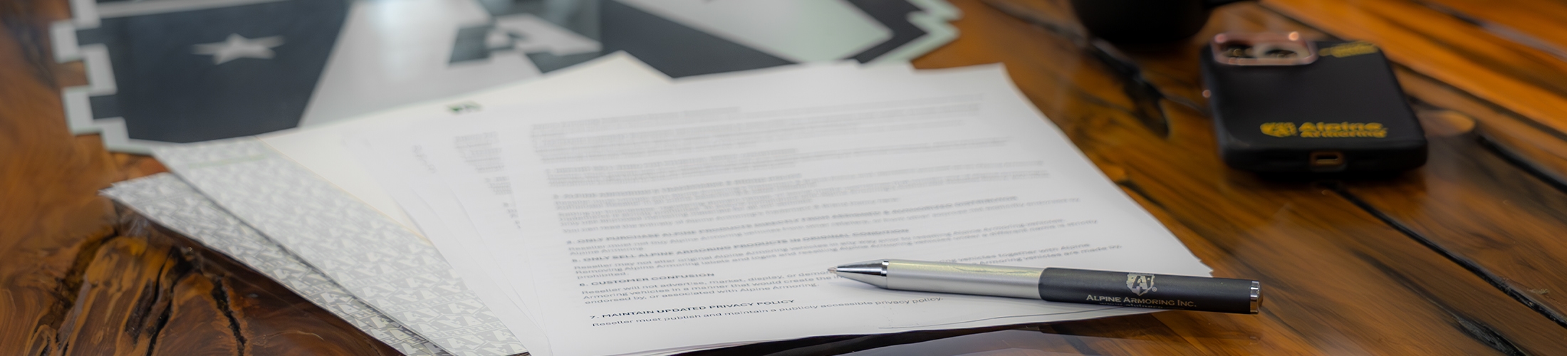 A pen rests on top of a stack of documents on a wooden table. Additionally, there are a smartphone and a large envelope nearby.