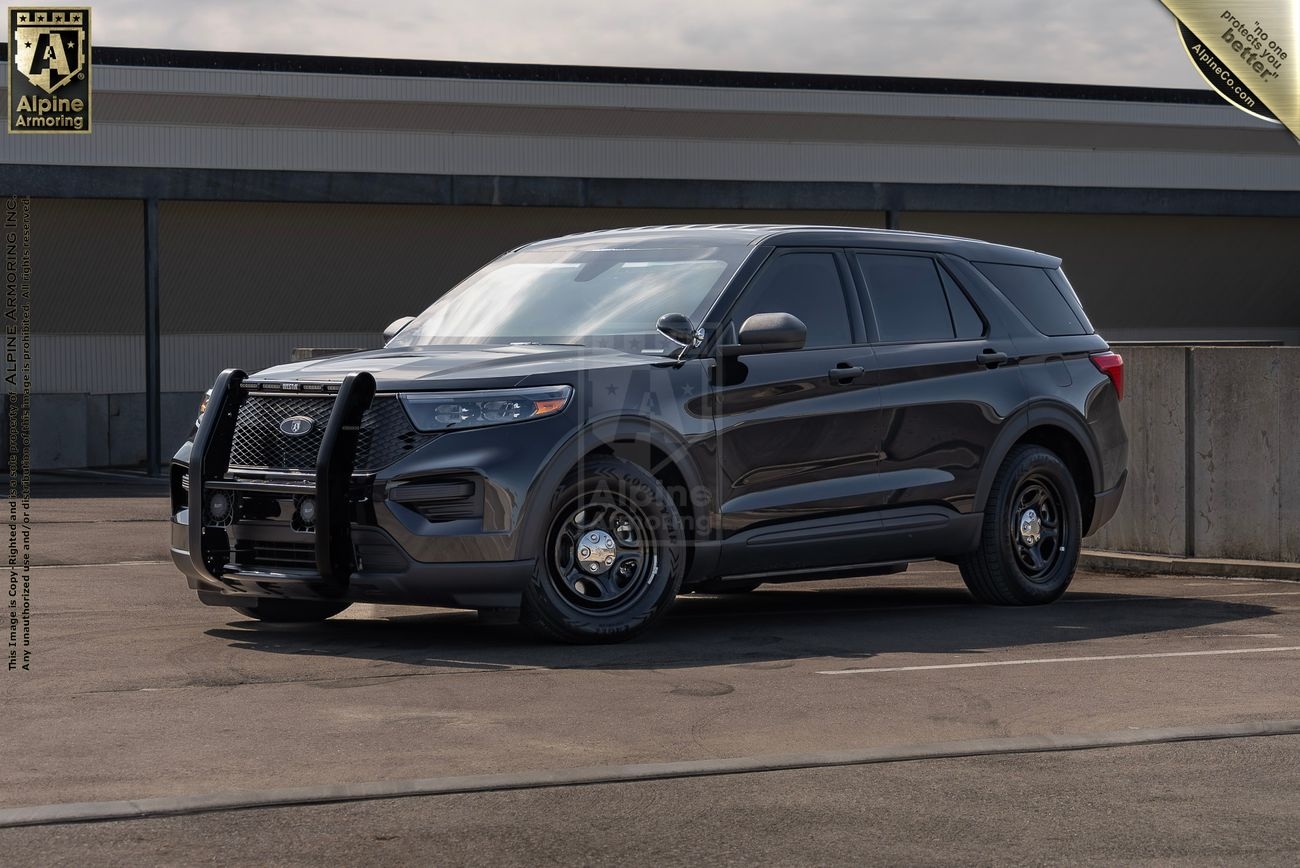 A black Ford Explorer Police Protection Vehicle is parked in a lot with barriers on a clear day. The vehicle has a front push bumper and dark tinted windows.