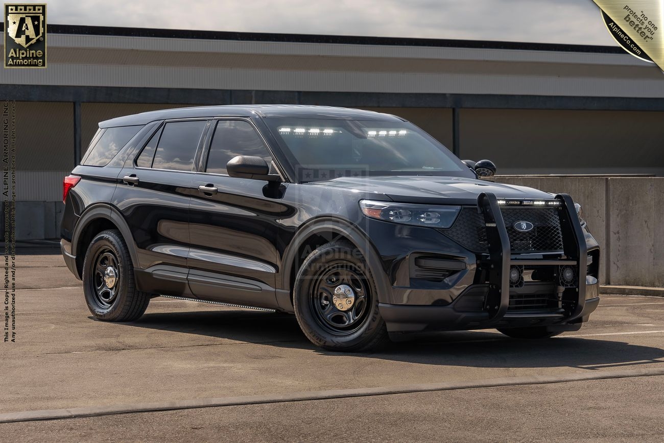 A black Ford Explorer police protection vehicle is parked on a concrete surface in front of a building. The vehicle has a bull bar on the front bumper and police lights on the roof. Logos and text are superimposed on the photo.