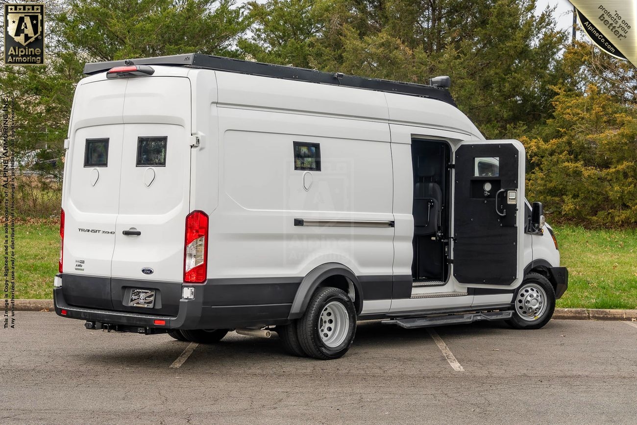 A white Ford Transit Pointer van with rear and side windows is parked outside. The side door is open, revealing the interior. Trees and a parking lot are visible in the background.