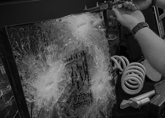 A person uses calipers to measure the diameter of bullet holes in a shattered glass surface, which is part of a computer tower. The glass is heavily damaged with spider-web cracks radiating from several impact points. 