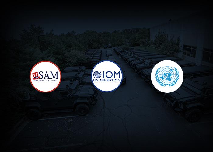  Three logos against a dark background of parked military vehicles: SAM logo (shield with three red lines), IOM UN Migration logo (blue circle with globe and human figures), UN logo.