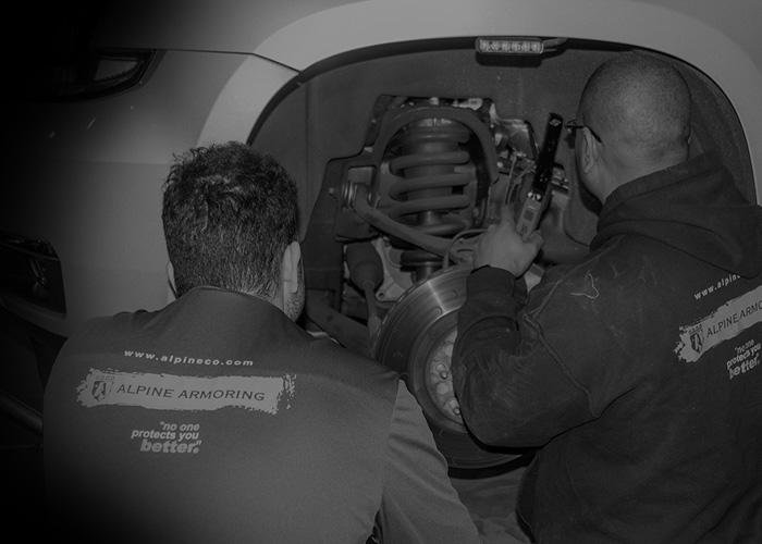 Two mechanics working on the wheel area of a vehicle. Both are wearing jackets with "Alpine Armoring" printed on the back.