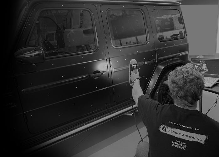 Amn Alpine Armoring employee uses a handheld device to inspect a black armored vehicle from Alpine Armoring, which has visible rivets along its side.