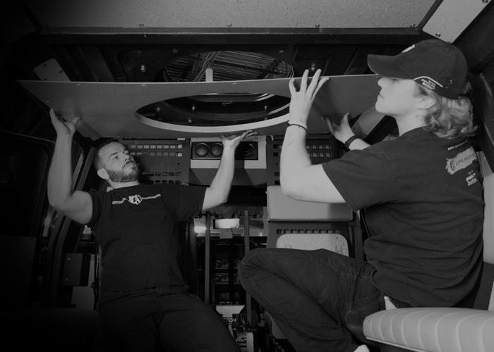 Two Alpine Armoring employees are installing a large panel in a mechanical or technical room. Both are using both hands to position the panel overhead. They appear to be working inside a vehicle or equipment enclosure.