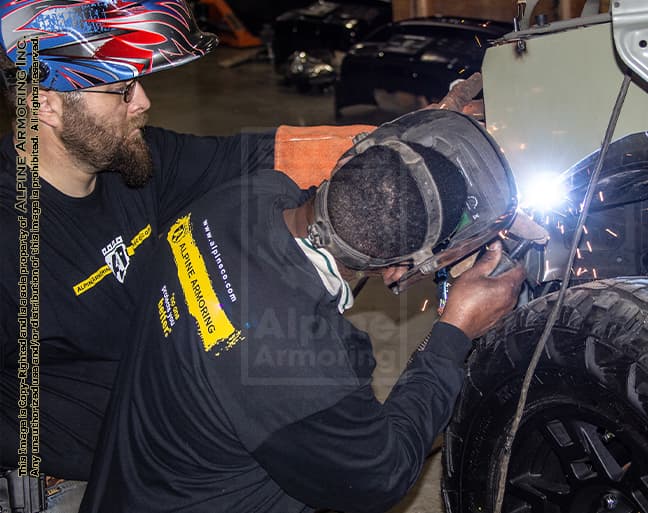 Two technicians at Alpine Armoring welding and assembling parts on an armored vehicle. Both are wearing protective gear, including helmets and gloves, highlighting the precision and safety measures in the manufacturing process.