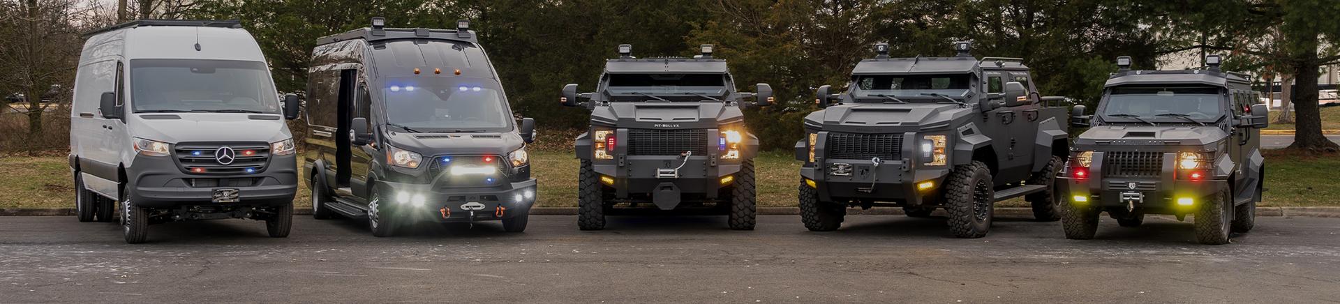 Five armored vehicles from Alpine Armoring on a paved surface at dusk: two vans on the left and three large, heavily armored trucks on the right, all with lights illuminated.