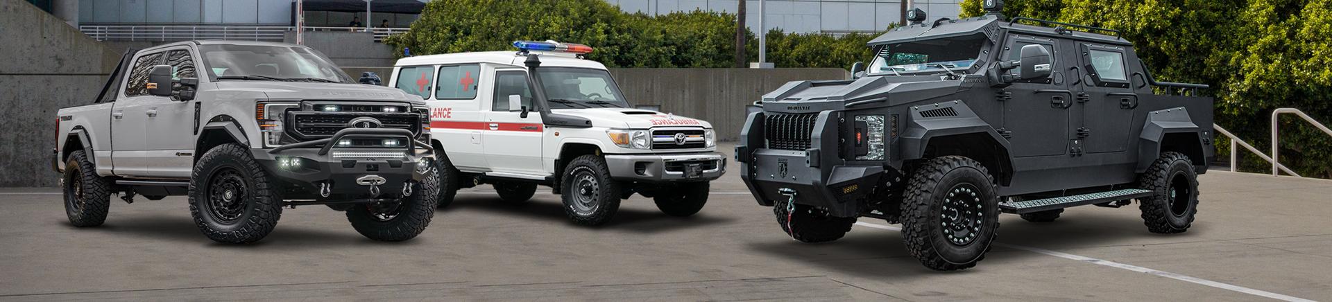  Three Alpine Armoring vehicles on a lot: a gray pickup truck with off-road mods, an emergency response vehicle with red and blue lights, and a black armored SUV with military features.