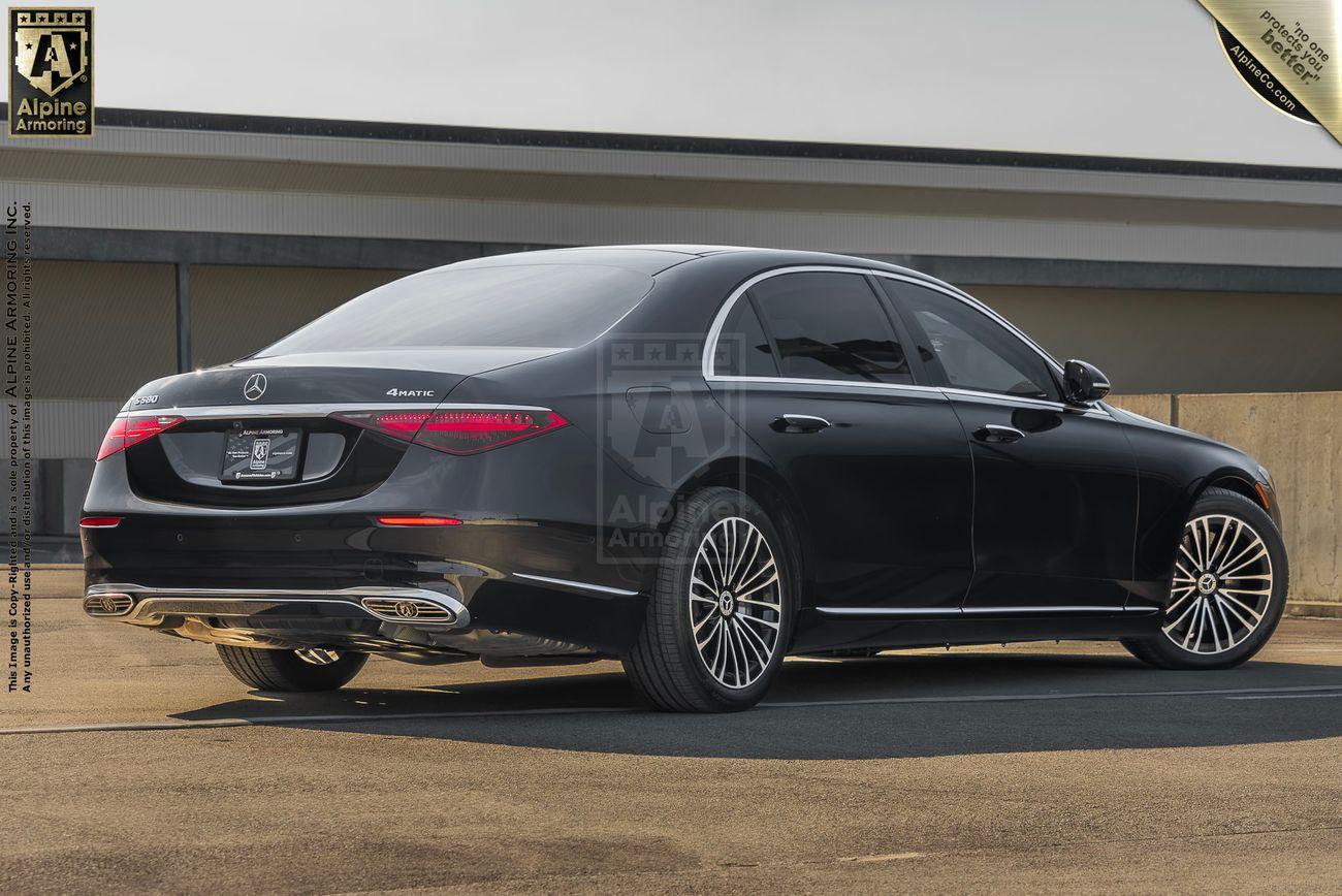 A black, armored Mercedes-Benz S580 sedan is parked on a concrete surface next to a building. A "Alpine Armoring" logo and details are imprinted on the image.