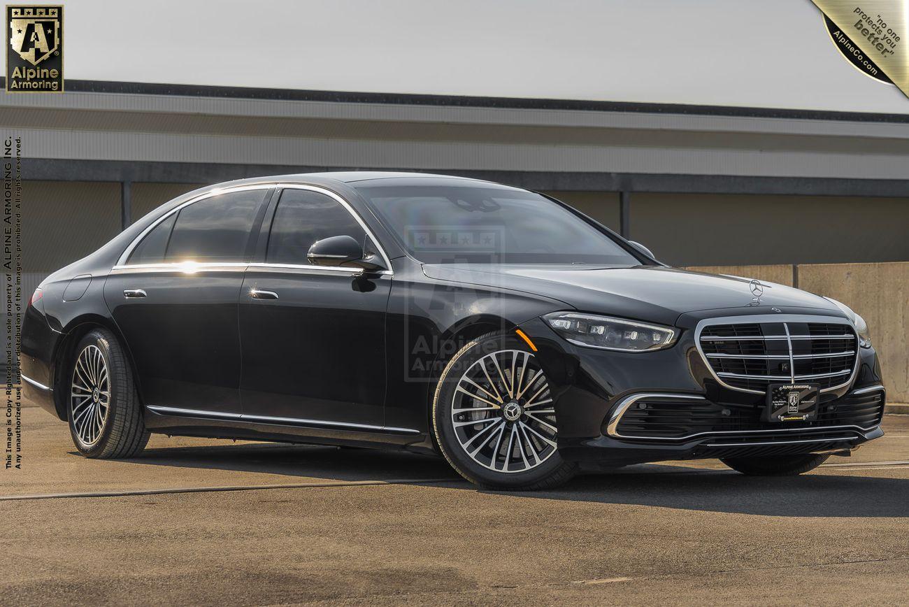 A black, armored Mercedes-Benz S580 luxury sedan parked in front of a modern building, featuring a large front grille, distinctive headlights, and polished wheels. An Alpine Armor plate and logo are visible on the top left.