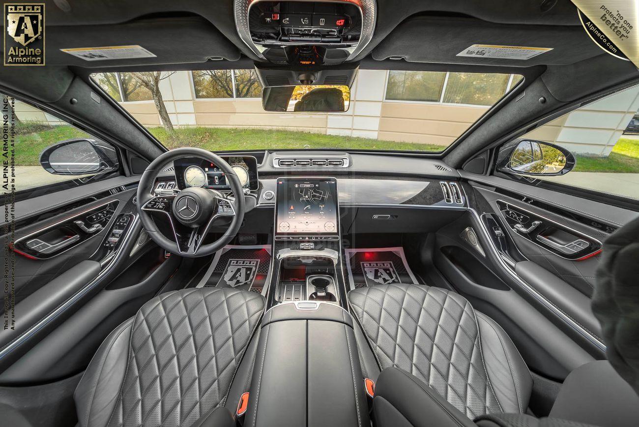 A luxurious black Mercedes-Benz S580 sedan interior with black quilted leather seats, a large central touchscreen display, and a sleek dashboard. Views through the car windows show a building and greenery outside.