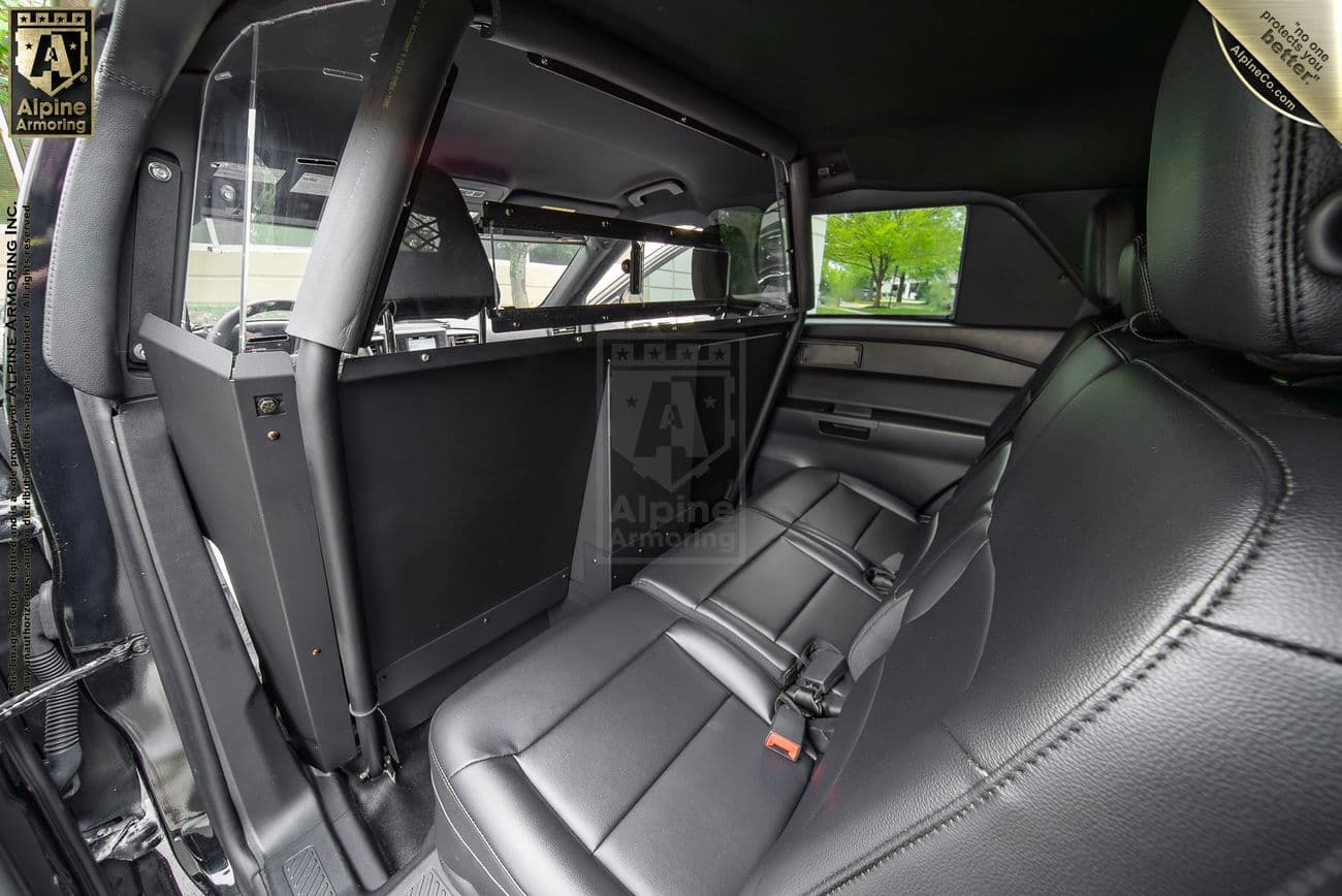 Interior of a police protection vehicle (PPV) showing the back seat with a protective partition and secure seating. The vehicle features black leather seats and a transparent screen dividing the front and rear areas.