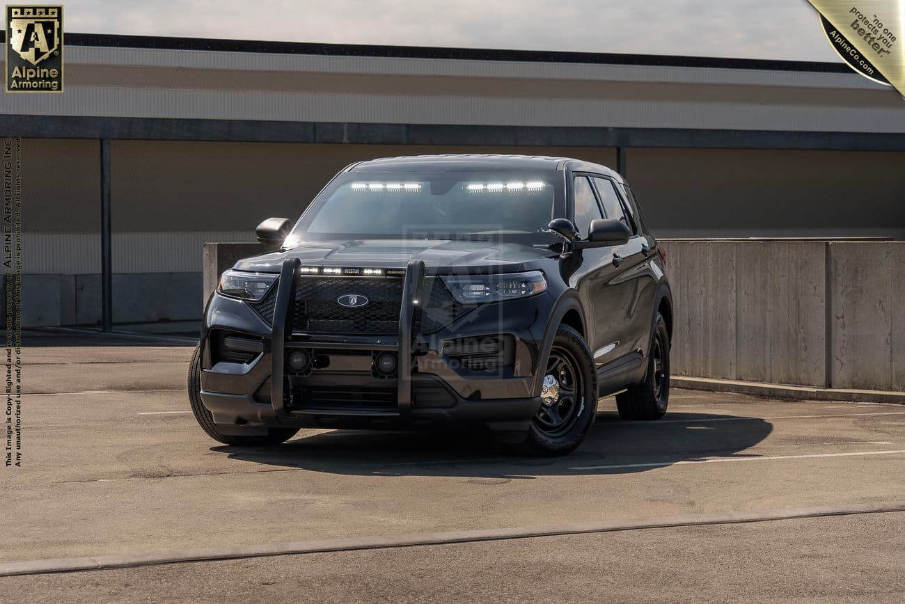 A black Ford Explorer PPV is parked in an open lot next to a concrete barrier under a partly cloudy sky. The vehicle is equipped with front push bars and roof-mounted lights.