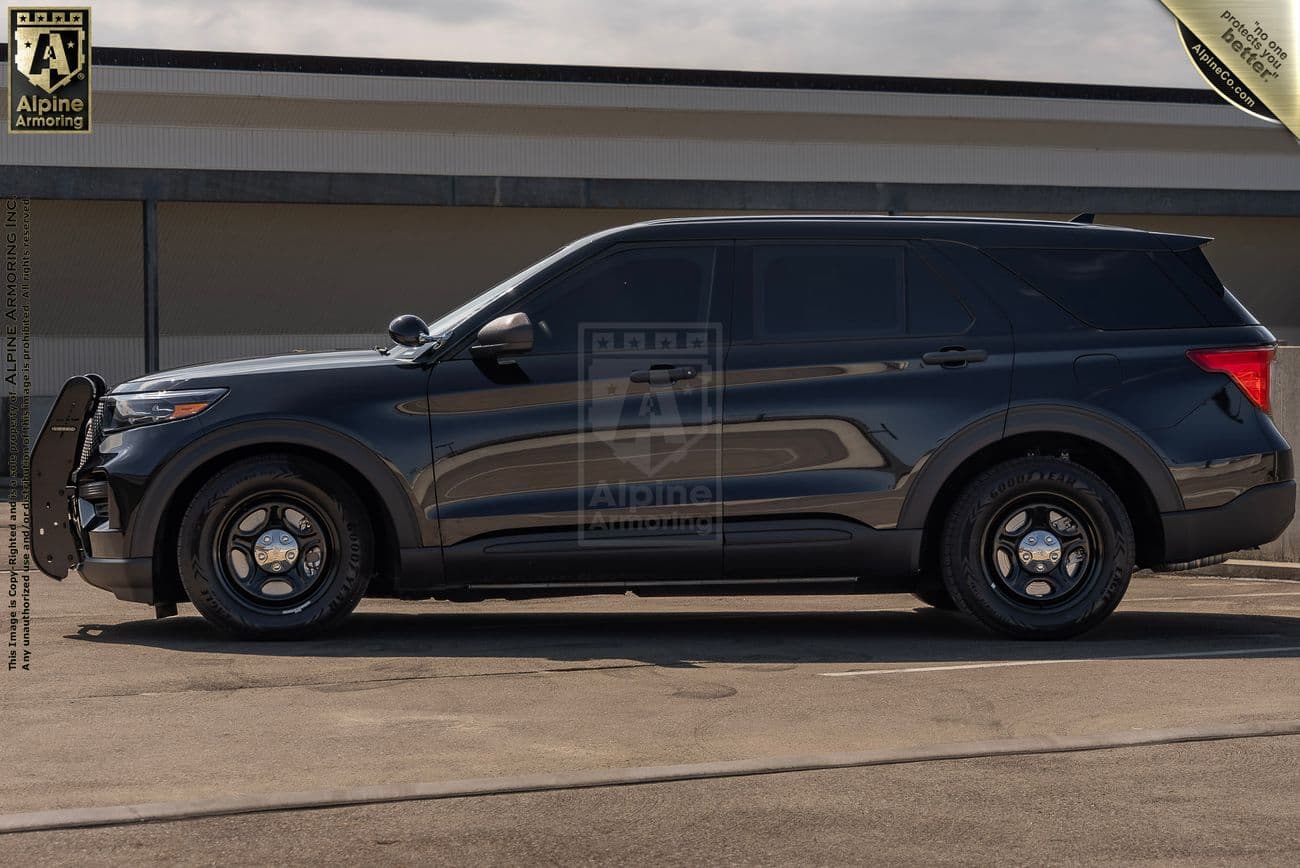 A large, black Ford Explorer PPV with black rims parked in front of a building. The vehicle has tinted windows and police modifications, including a push bumper on the front.