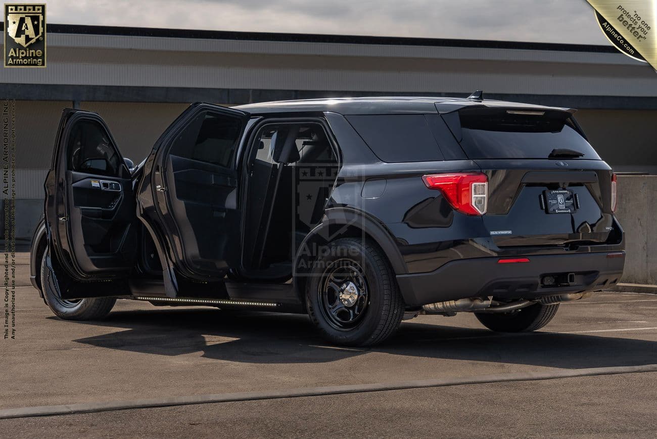 A black Ford Explorer PPV (Police Protection Vehicle) with doors open, showcasing the interior. The vehicle appears to be an armored model from Alpine Armoring. The SUV is parked on a concrete surface with a building in the background.