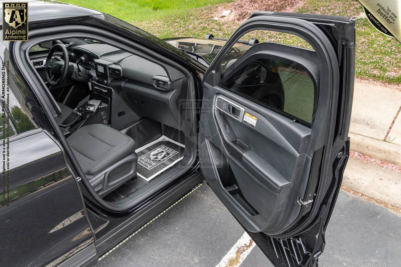 The interior and open front door of a black Ford Explorer Police Pritection Vehicle (PPV). The driver's seat and steering wheel are visible. A protective cover is on the floor mat, and the window is partially rolled down.