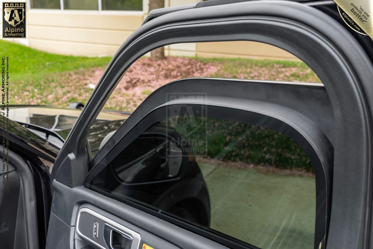 Close-up of a partially opened car door with a bulletproof glass window installed. The background includes a building, a window, and some grass.