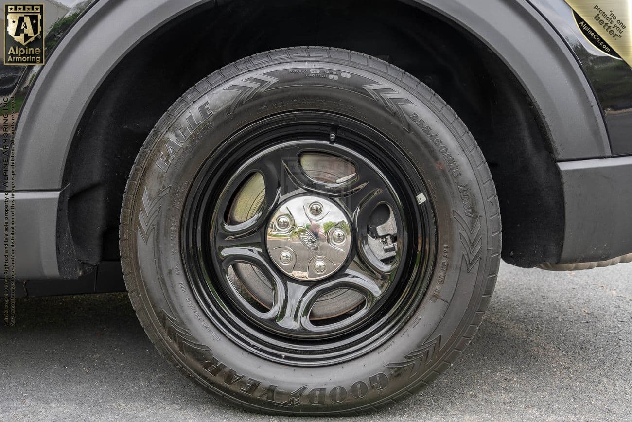 Close-up of a black car wheel and tire with a shiny hubcap, partially visible dark vehicle body, and “Alpine Armor’ing” logo on the left side of the image.