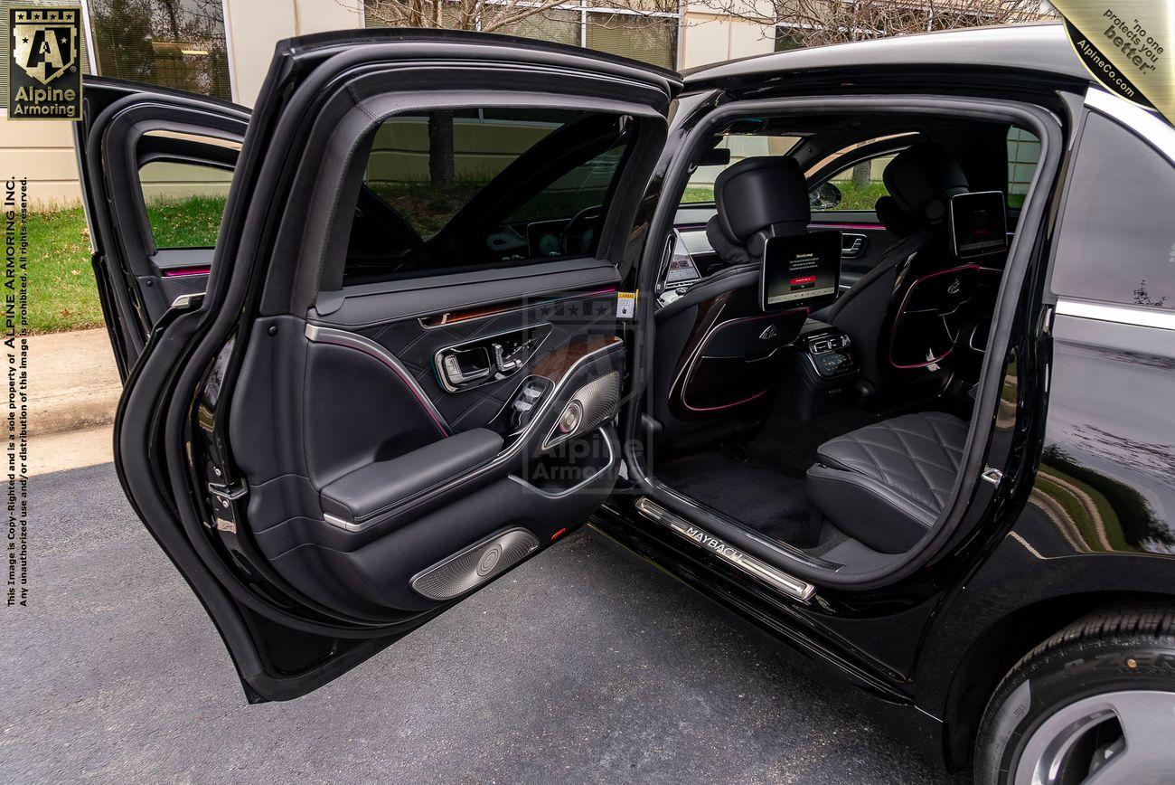 Interior view of a luxurious Mercedes-Benz S 580 Maybach sedan with open rear passenger doors showcasing plush seating, multiple screens, and premium finishes. The car has black leather seats with a quilted pattern and ambient lighting.