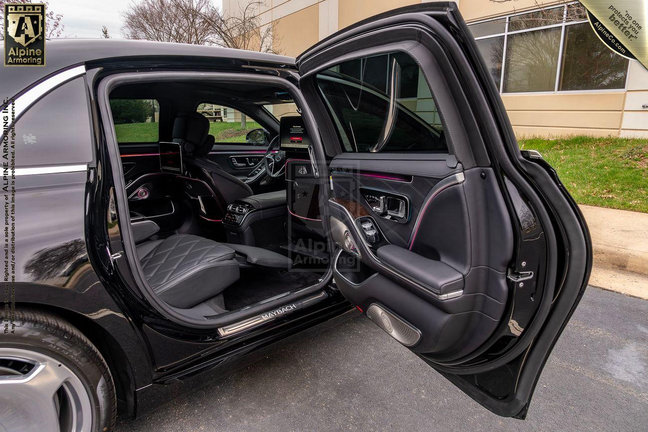 An open black Mercedes-Benz S 580 Maybach sedan door reveals a luxurious black leather interior with red accents. The front and back seats are visible, and the car is parked outside near a building with a glass window.