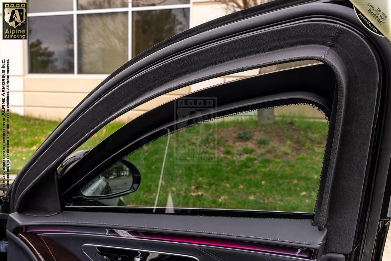 An open armored Mercedes-Benz S 580 Maybach sedan door showing the interior of a vehicle with tinted windows and a green lawn visible through the bulletproof glass door window.