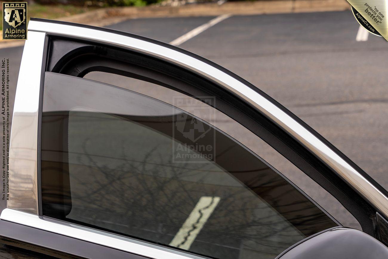 Close-up side view of an armored Mercedes-Benz S 580 Maybach window with bulletproof glass, partially open, showing metallic trim and tinted glass. Alpine Armoring logo and text are visible on the window.