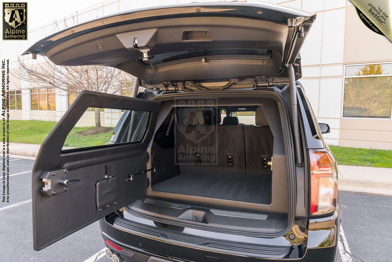 A black armored Chevrolet Suburban LT SUV with its rear hatch and the left-side rear window open, showing a spacious, empty cargo area. .