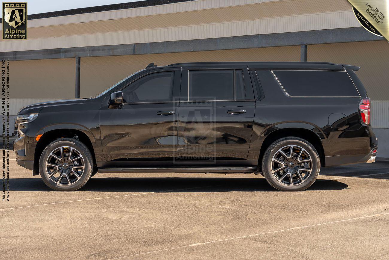 Side view of a black armored Chevrolet Suburban LT SUV with dark-tinted windows, parked on a concrete surface in front of a beige building.