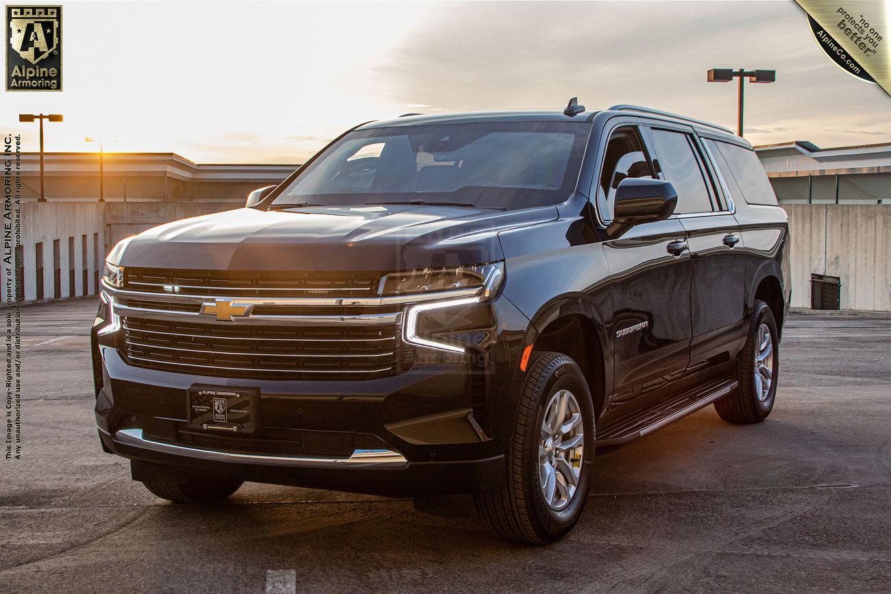 A black, bulletproof Chevrolet Suburban LT SUV is parked in an outdoor parking lot at sunset, with a building and streetlights visible in the background.