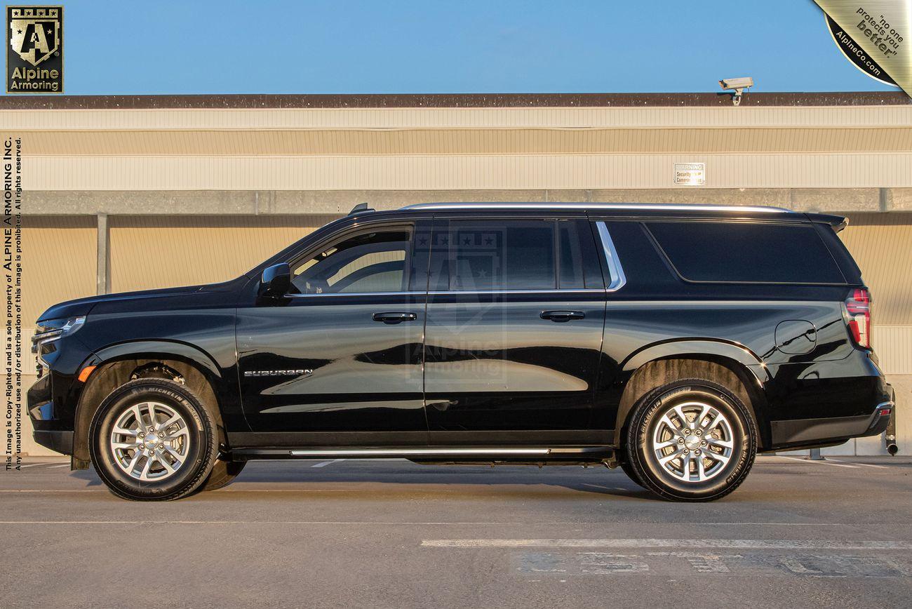 A black armored Chevrolet Suburban LT SUV is parked in a lot, viewed from the side. The vehicle has four doors and tinted windows. The background features a beige building and a clear sky.