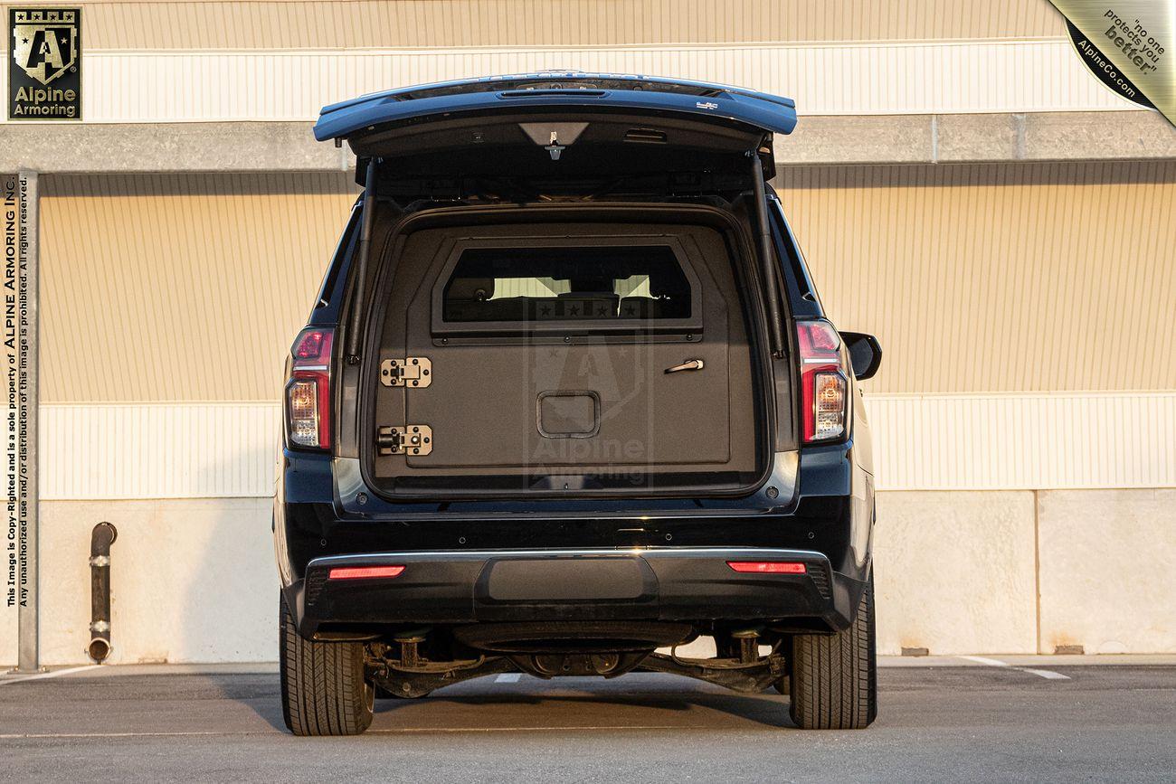 An armored Chevrolet Suburban LT SUV with its rear door open, showing a secure compartment inside. The exterior is black with logos for Alpine Armoring on the sides.