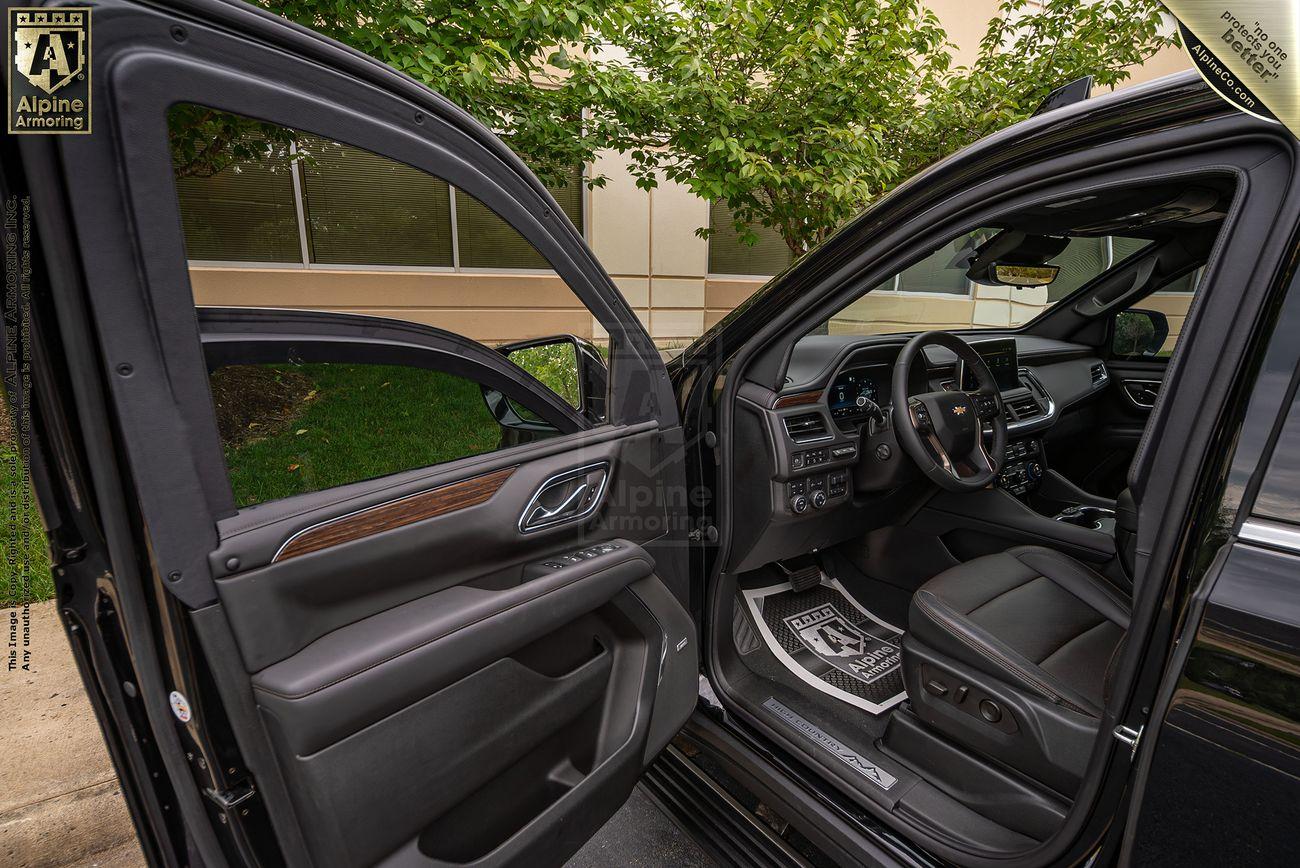 Interior view of a black armored Chevrolet Suburban LT SUV with the driver's side door open, displaying leather seats and the dashboard. Trees and a building are visible in the background.