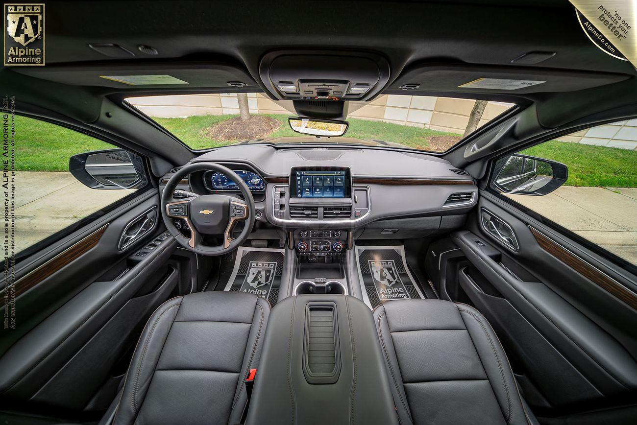 An interior view of an armored Chevrolet Suburban LT vehicle featuring a central touchscreen, black leather seats, and various control buttons on the dashboard. The logo "Alpine Armoring" is visible on the floor mats and the top corners.