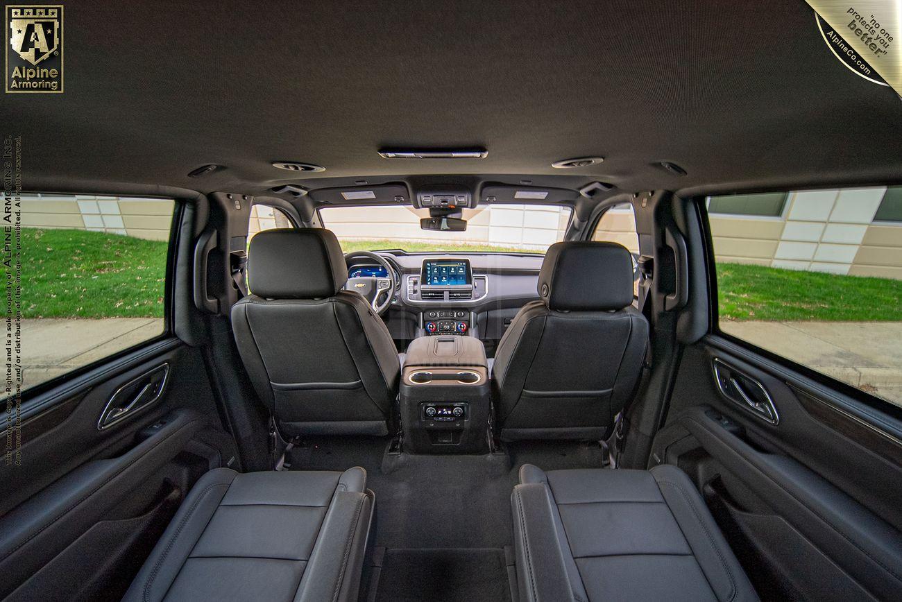 Interior view of a black bulletproof armored Chevrolet Suburban LT SUVshowing the dashboard, front and second-row seats, and rear windows, with a visible Alpine logo on the upper left. The vehicle is parked outside near a building.