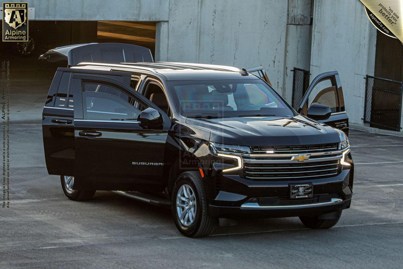 A black Chevrolet Suburban LT SUV with its doors open is parked in an outdoor setting. The vehicle features the emblem of Alpine Armoring.