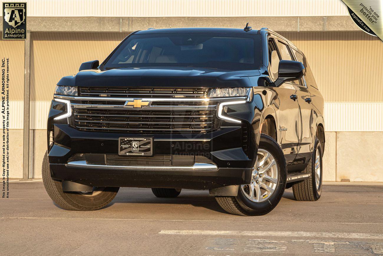 An armored black Chevrolet Suburban LT SUV is parked in an outdoor parking lot at sunset, with a building and streetlights visible in the background.