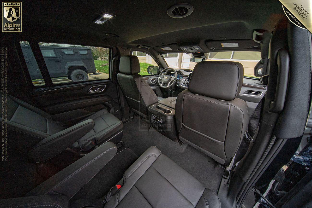 Interior view of a Chevrolet Suburban LT SUV showcasing black leather seats, center console space, and overhead lighting. An Alpine Armoring logo is visible in the top left corner.