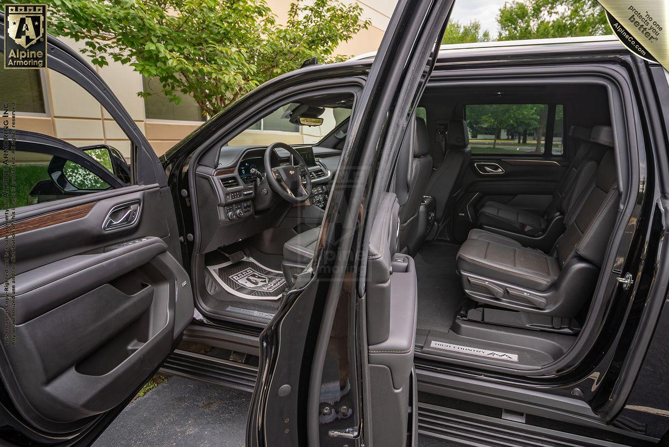 Interior view of a black armored Chevrolet Suburban LT SUV with the driver's door and rear passenger door open, showing the front and rear seats, dashboard, and steering wheel. Trees and a building are visible outside.