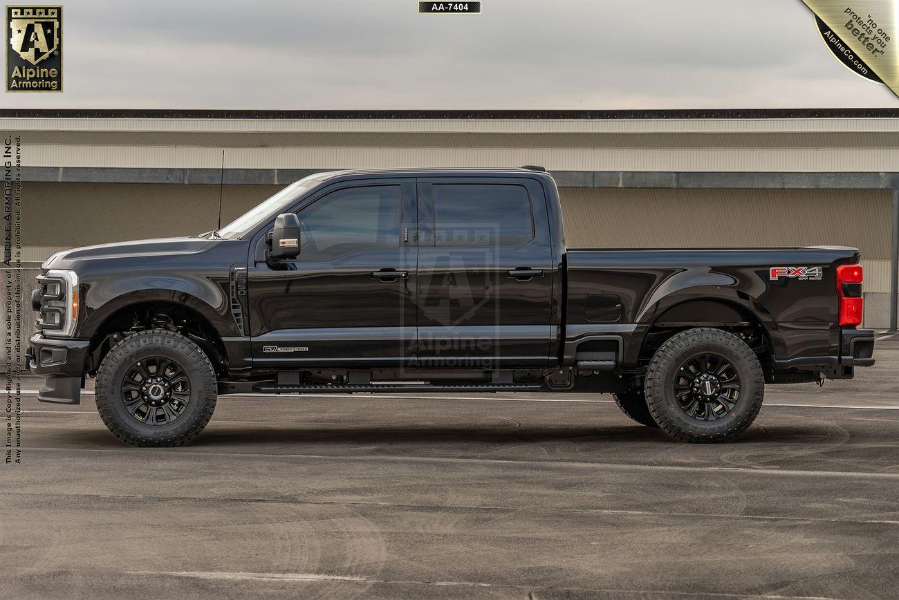 Side view of a black bulletproof Ford Super Duty F-350 Lariat pickup truck parked on a concrete surface in front of a building. The truck has large off-road tires and a crew cab configuration.