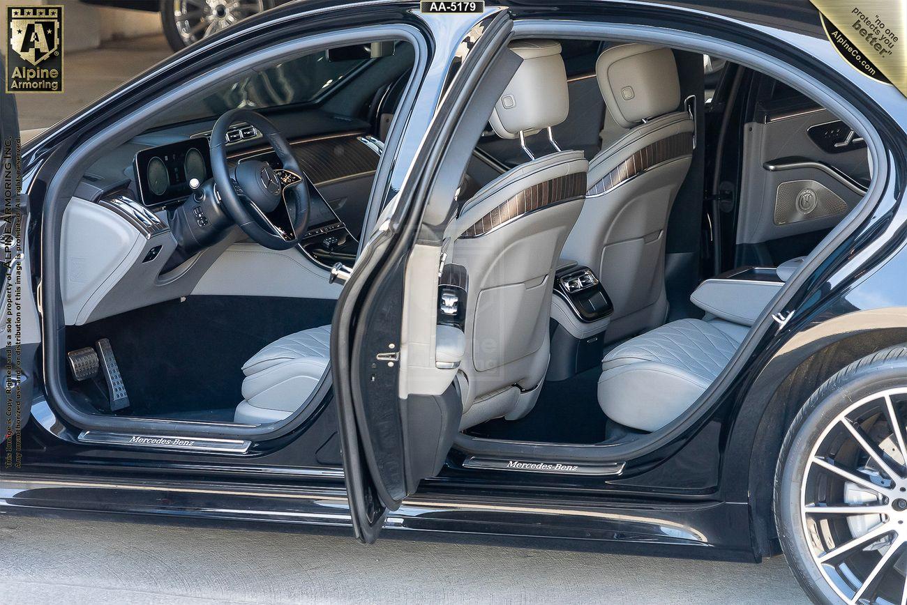 Interior view of a armored Mercedes S580 S-Class 4MATIC sedan with open rear passenger door showing leather seats, wood trim, and a center console with controls. The car is parked in a garage.