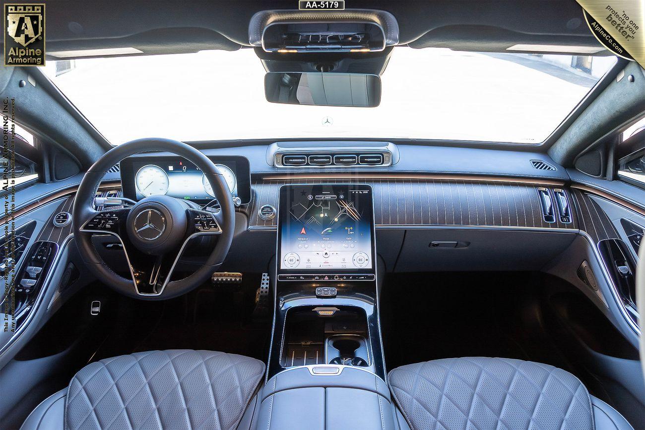 Interior of a black armored Mercedes-Benz S580 S-Class 4MATIC sedan showing the dashboard, steering wheel, large central touchscreen, quilted leather seats, and wood trim accents. The badge on the dashboard indicates Alpine Armoring.