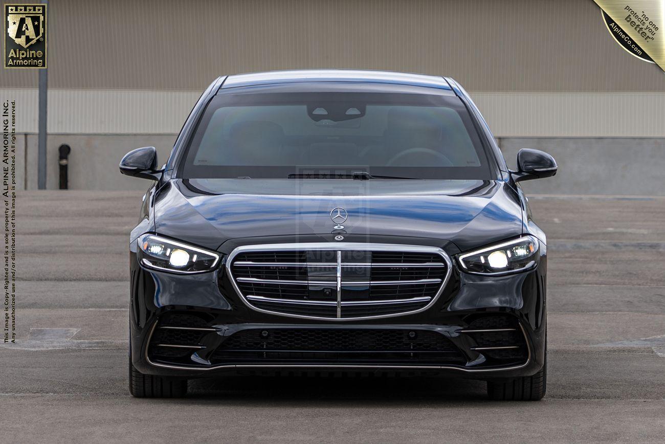 Front view of a luxurious black Mercedes S580 sedan parked on a paved surface, with Alpine Armoring branding visible on the vehicle and in the background.