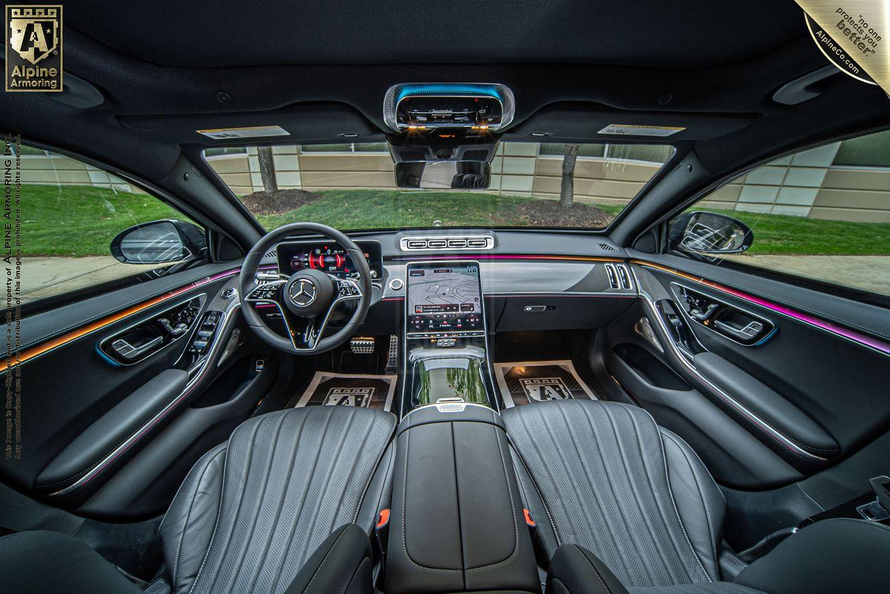 A wide-angle view of the front interior of a ln armored black Mercedes-Benz S580 sedan, showcasing black leather seats, a large touchscreen dashboard console, and a sunroof. The car has Alpine branding visible on the floor mats.