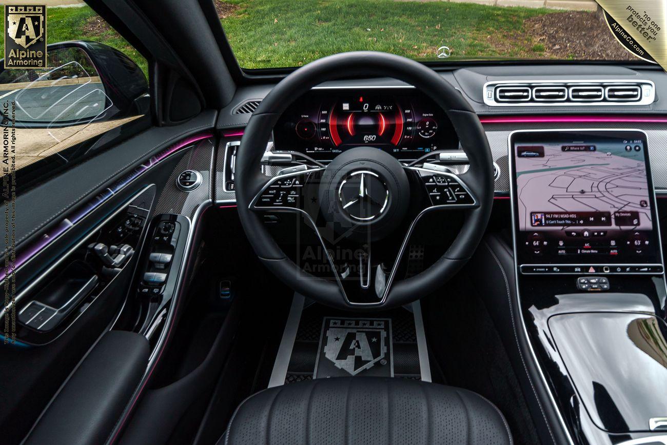 Interior of an armored black Mercedes-Benz S580 sedanshowcasing a steering wheel with a prominent emblem, digital dashboard displays, touchscreen navigation system, and ambient lighting. Exterior view through the windshield.