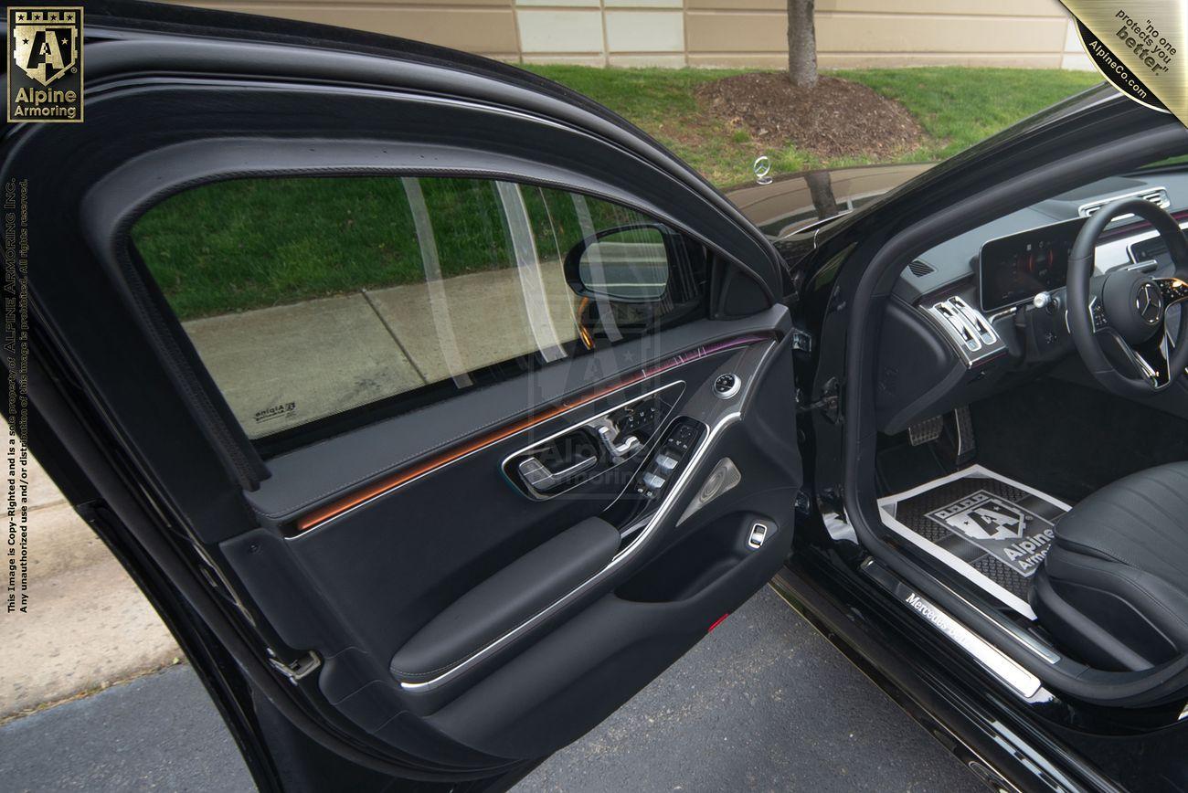 View of an open car door showing the interior of a black Mercedes-Benz S580 sedan with visible control buttons, bulletproof glass window, seat, and steering wheel. Outside, there's a sidewalk, grass, and a building wall.