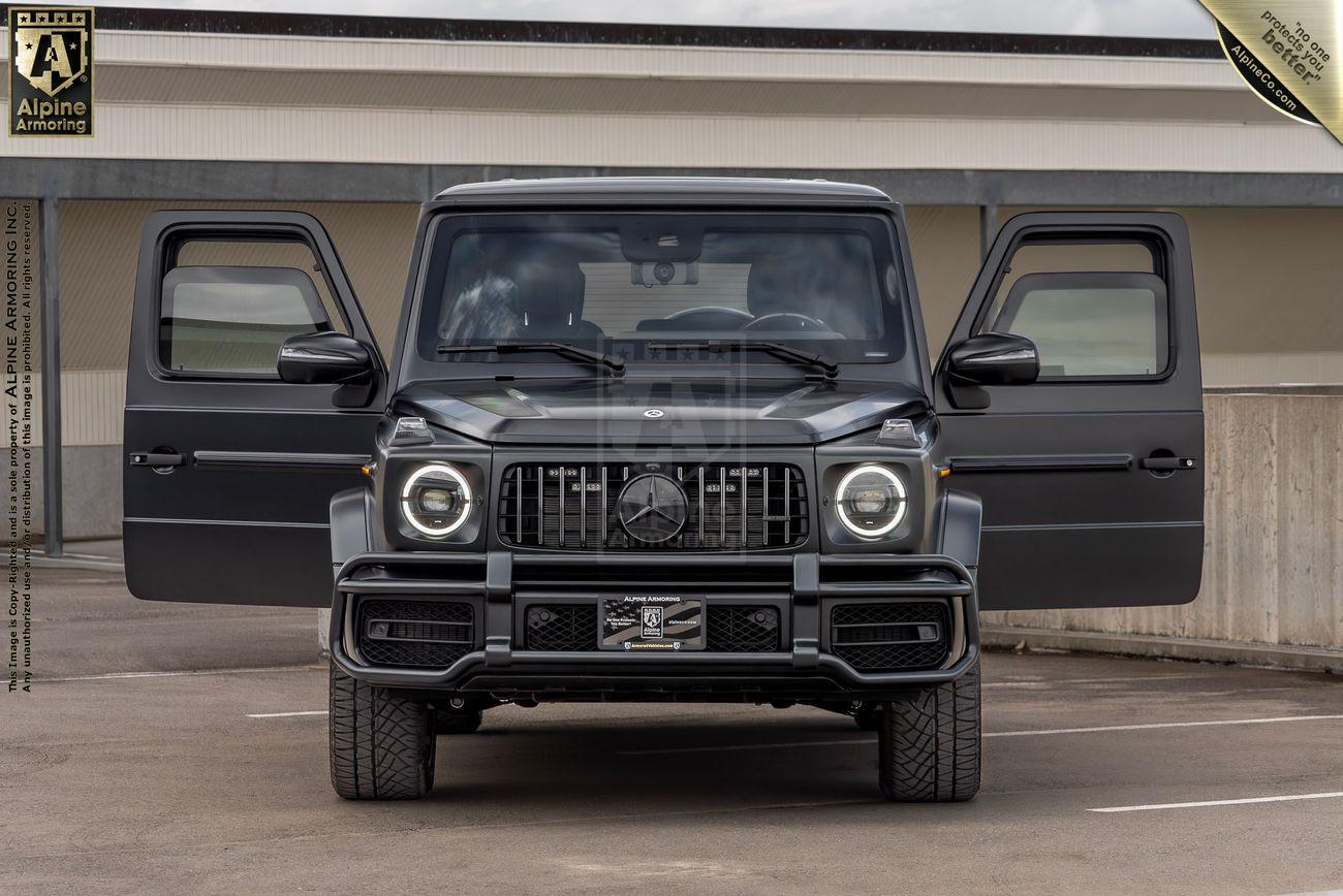 Front view of a dark-colored Mercedes-Benz G-Class SUV with its front doors open, parked outdoors on an asphalt surface with trees in the background.