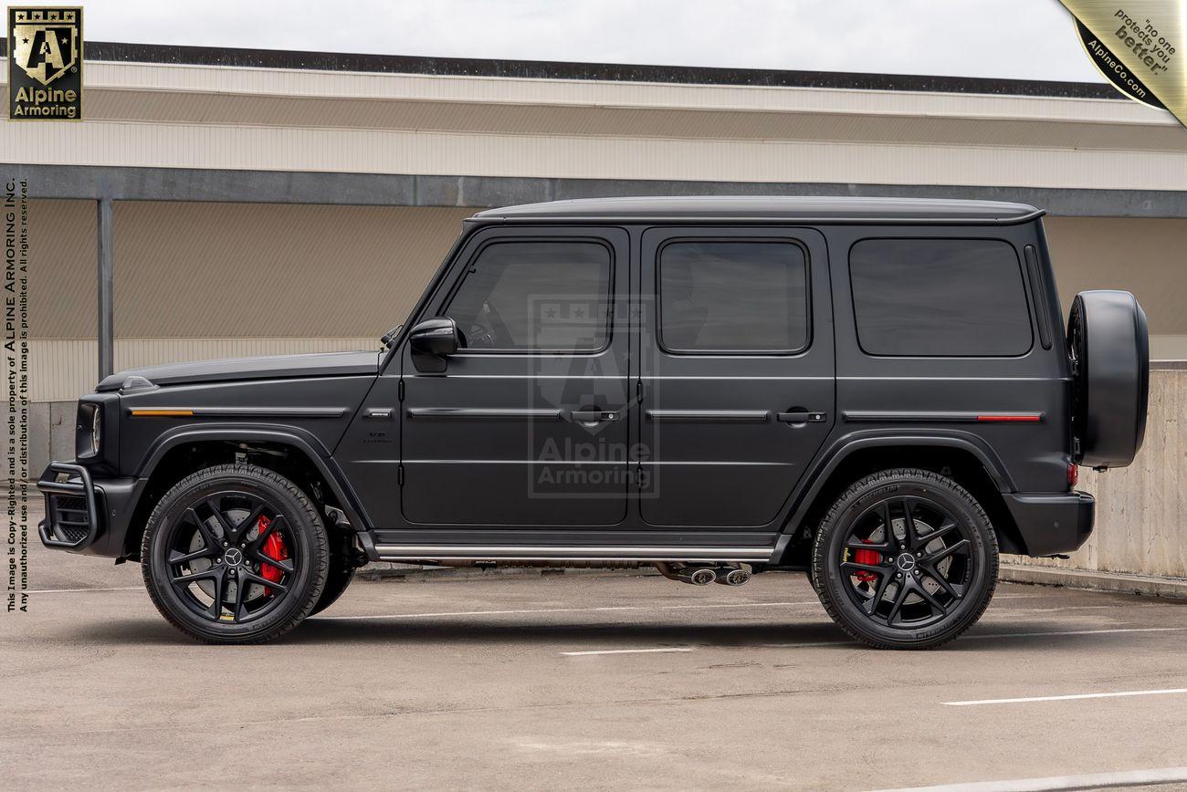 A black Mercedes-Benz AMG G-Class SUV is parked in a parking lot in front of autumn foliage. The vehicle has a matte finish and dark-tinted windows.