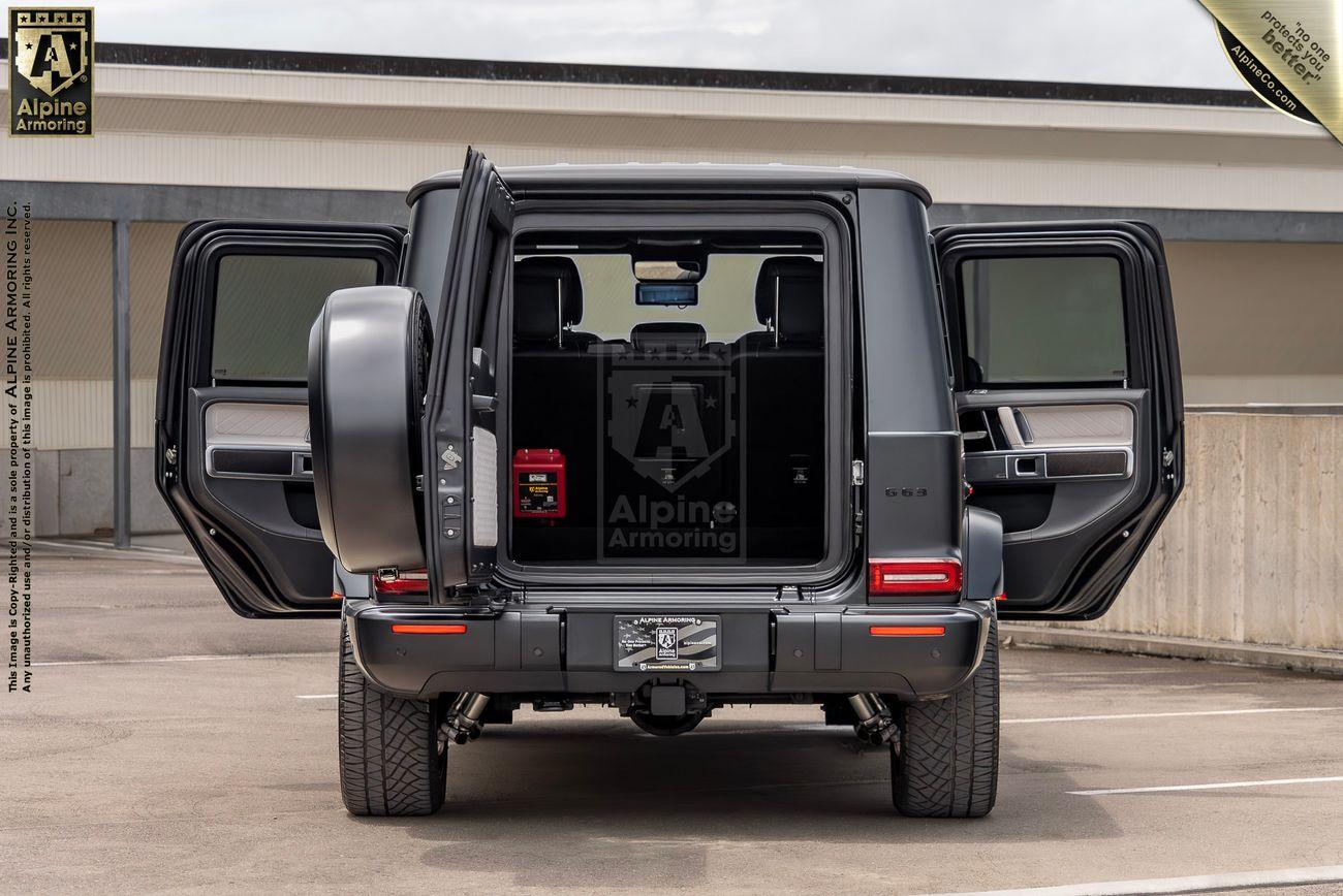 A rear view of a black Mercedes-Benz G-Class SUV with a visible Alpine Armoring logo on the tire cover, parked in a lot with trees in the background.