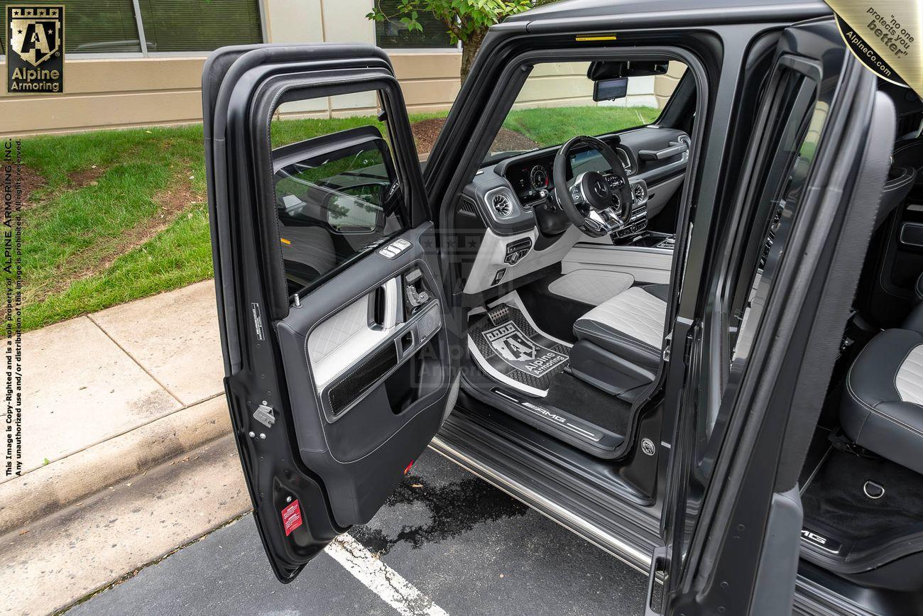 Close-up of a G-Class SUV's bulletproof window, partially rolled down, with a forested background and a company logo in the upper left corner.