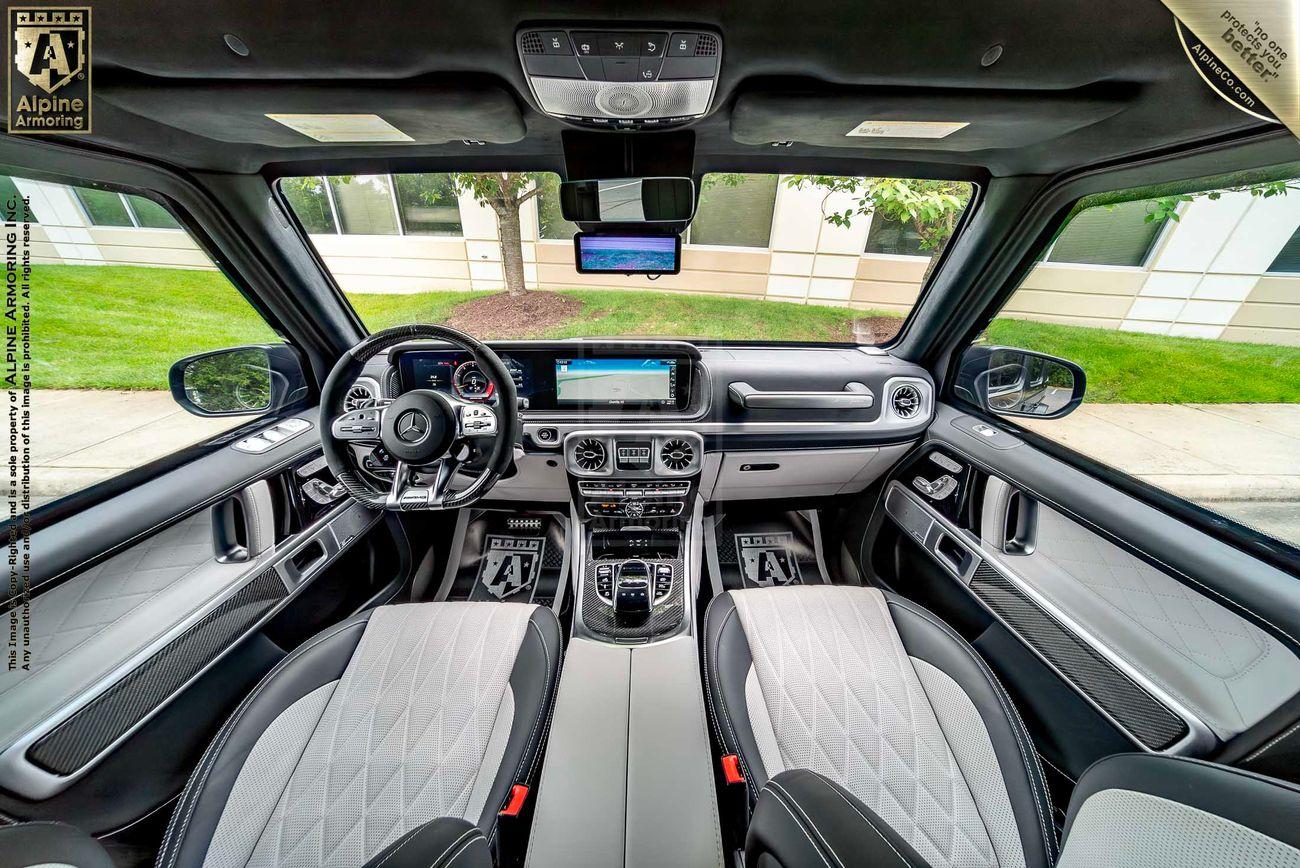 Interior of a luxury Mercedes SUV showing the dashboard, steering wheel, infotainment system, and two front seats with diamond-stitched upholstery and wooded area visible through the windshield.